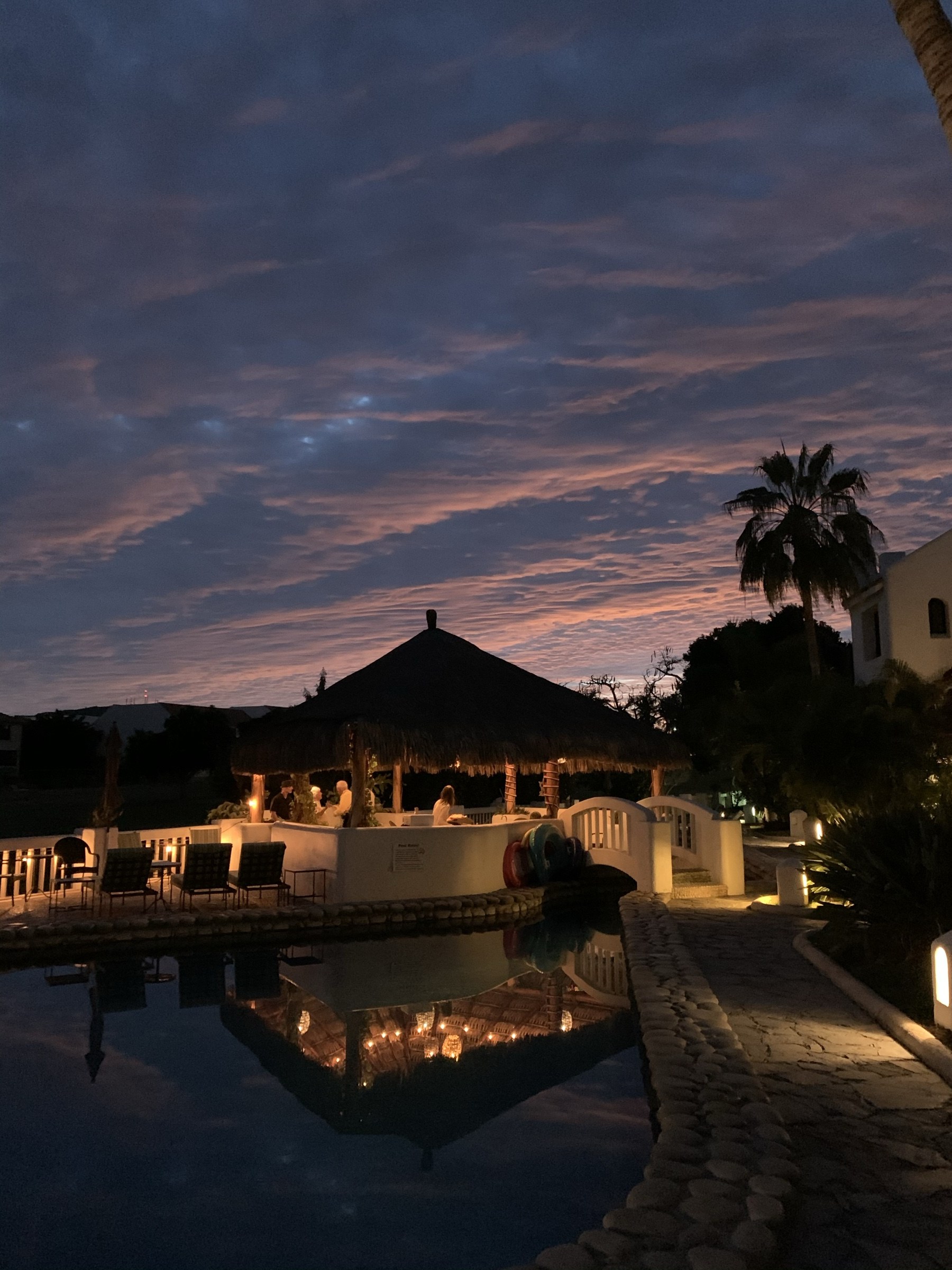 Evening pool scene with palapa, warm lights, and sunset sky